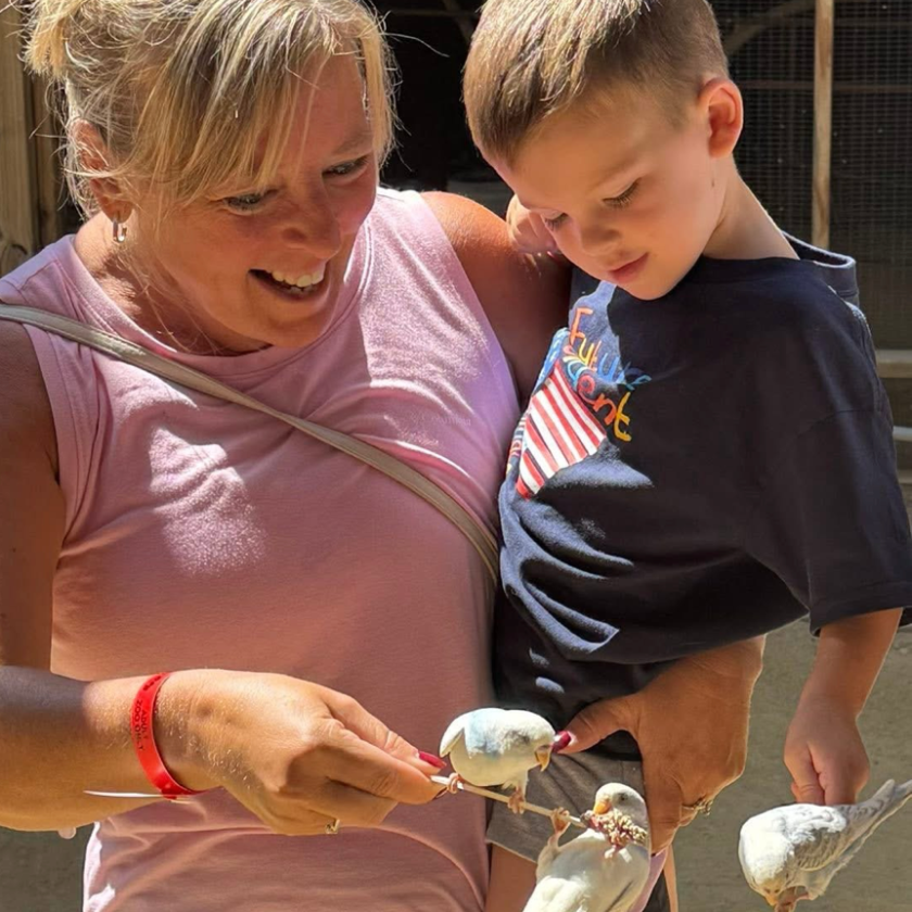 Woman and child feeding small white birds on sticks in a sunny outdoor setting.